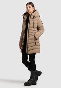 Beżowy, ocieplany płaszcz o średniej długości, z kapturem, zamkiem błyskawicznym z przodu i bocznymi kieszeniami. Nosi go na czarnym topie, w połączeniu z czarnymi butami wojskowymi.