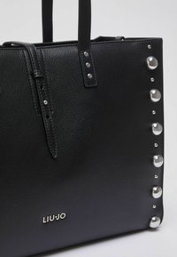 Sac cabas en cuir noir avec une surface texturée, des clous décoratifs argentés sur un côté, et un logo embossé en bas.