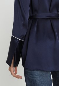 Blusa de satén azul marino con cintura ceñida, puños en contraste y detalle de ribete blanco en las mangas. Textura suave con forma ajustada.