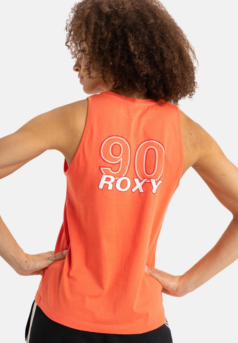 Femme aux cheveux bouclés portant un haut sans manches orange avec "90 ROXY" imprimé dans le dos, mains posées sur les hanches.