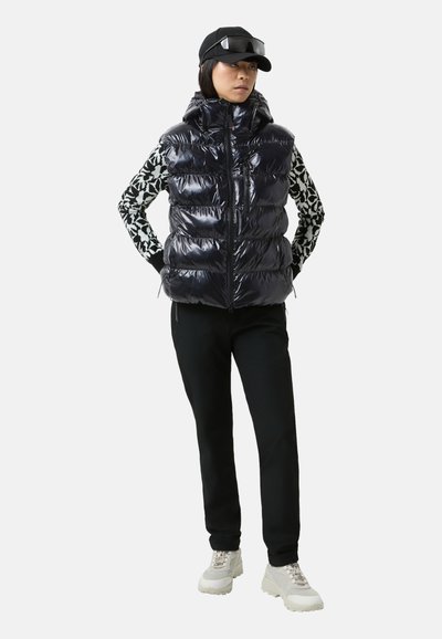 Błyszcząca czarna kamizelka puchowa z kapturem, zapinana na zamek, z bocznymi kieszeniami. Nosi się ją na wzorzystą bluzkę z długim rękawem, w połączeniu z czarnymi spodniami i sneakersami.