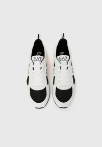 Sportlikud kingad valges ja mustas toonis, millel on võrgust pealmine osa, ümar ninaosa, paelad, bränditud keel ning aktsendid külgedel.