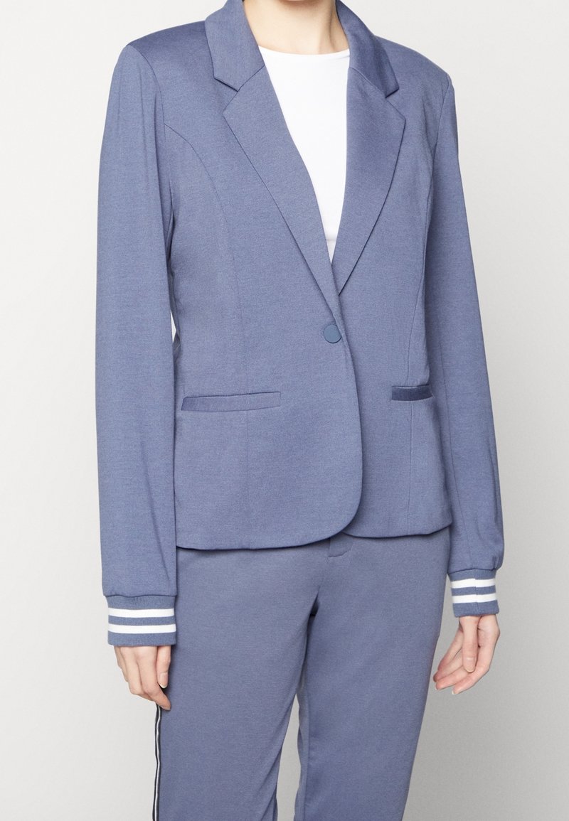 Femme portant un blazer bleu clair à un bouton, assorti d’un pantalon avec bandes latérales et une chemise blanche en dessous.