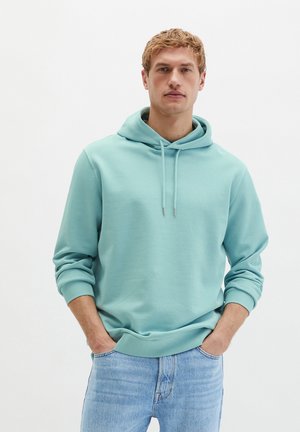 Blonder Mann, der einen hellblauen Hoodie und Jeans trägt, steht mit den Händen in den Taschen vor einem schlichten weißen Hintergrund.