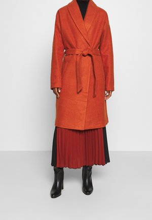 Personne portant un manteau en laine orange rouille ceinturé, par-dessus une jupe plissée noire et rouge, avec des bottines noires à talons sur fond blanc.