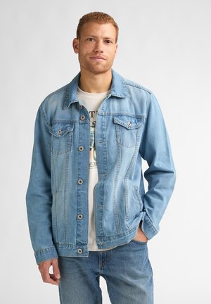 Hellblaue Jeansjacke mit Knopfleiste, zwei Brusttaschen und kontrastierenden Nähten. Das Model trägt darunter ein weißes T-Shirt mit Grafikdruck.