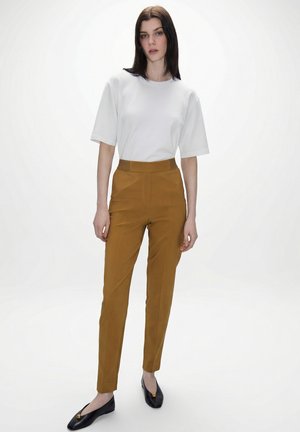 Frau mit glattem, dunklem Haar, die ein weißes Kurzarmshirt, senffarbene, hoch taillierte Hosen und schwarze Ballerinas mit goldenen Akzenten trägt.