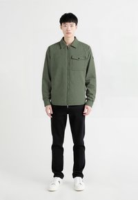 Olivengrøn lynlåsjakke med tekstureret stof, der har en brystlomme og sort knap, båret over sorte bukser og hvide sneakers.
