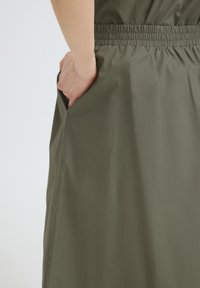 Main insérée dans la poche latérale d'une jupe vert olive avec une ceinture élastique froncée, sur un fond uni.