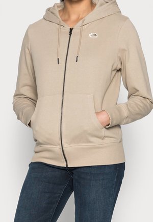 Beige hoodie met ritssluiting, trekkoorden en voorzakken, gedragen met donkerblauwe spijkerbroek, klein The North Face-logo op de borst.