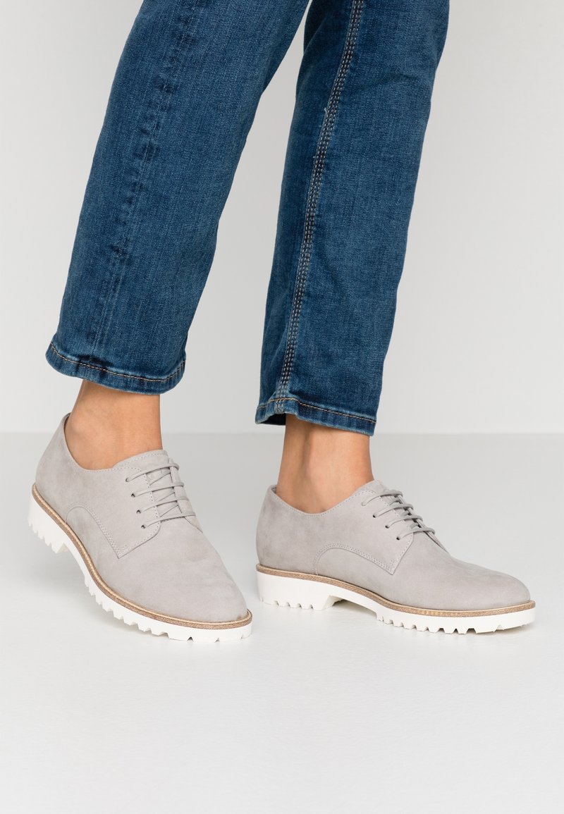 Personne portant un jean bleu et des chaussures en daim gris clair à lacets avec des semelles blanches texturées, debout sur un sol gris clair.