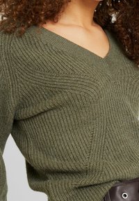 Kobieta ubrana w teksturowany oliwkowy sweter w serek oraz ciemno-brązowy skórzany pasek, z częściowo widocznymi kręconymi włosami.