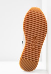 Semelle extérieure de la basket Lacoste, dotée d'une semelle en caoutchouc gum texturée avec des rainures horizontales et un logo en relief, contrastant avec une tige blanche.