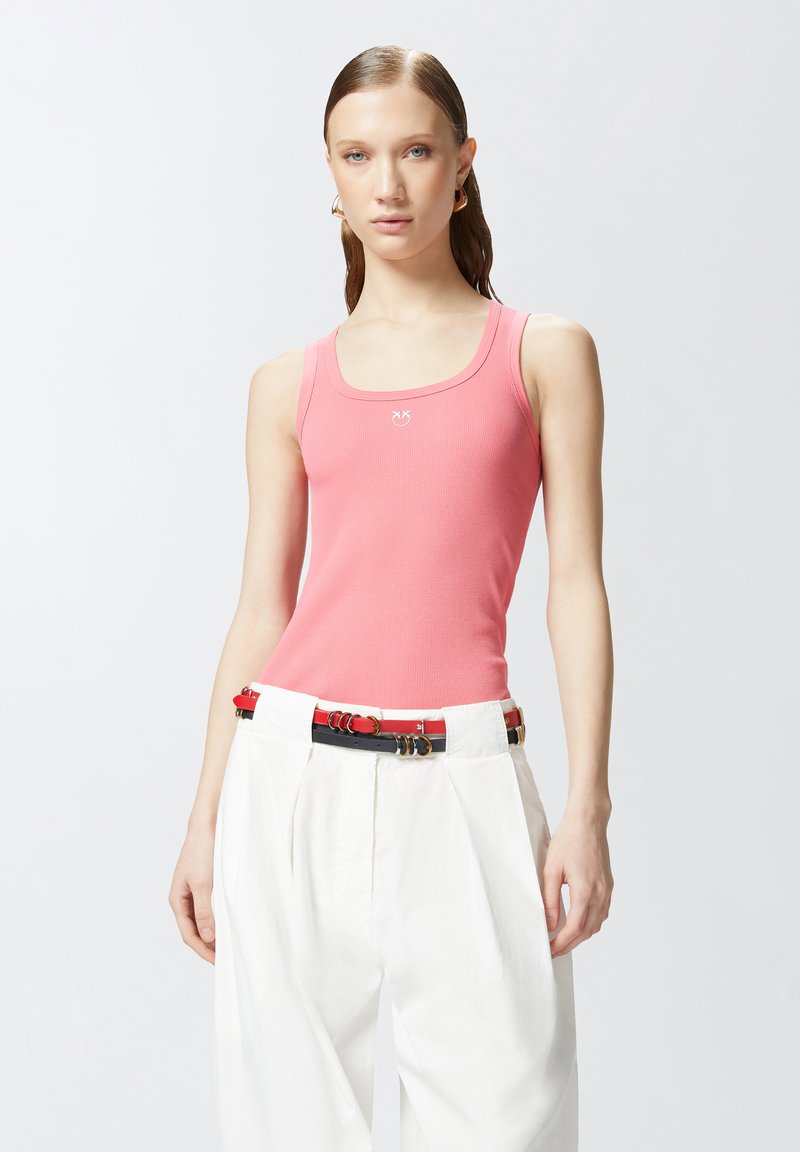 Ružičasta rebrasta majica bez rukava s izrezom u obliku suknje i malim logom. Uparena s hlačama visokog struka u bijeloj boji i šarenim pojasom.