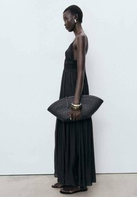 Robe maxi noire avec un corsage ajusté et une jupe fluide. Les accessoires comprennent un sac à main en osier et des bracelets jonc en or.