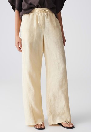 Frau trägt lockere, hoch taillierte, cremefarbene Leinenhose mit Tunnelzugbund und schwarze Zehensandalen, steht vor weißem Hintergrund.