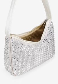 Bolso sintético en beige claro con tachuelas plateadas. Tiene un cierre de cremallera, un bolsillo interior y una correa de tela texturizada para llevar.