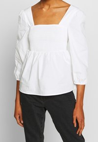 Vit peplumblus med fyrkantig halsringning, texturerad smockad byst och puffärmar, tillverkad av lätt material med elastiska manschetter.
