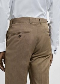 Pantaloni cu carouri maronii, cu o textură netedă, având două buzunare la spate cu închidere prin buton. Purtați cu o cămașă albă.