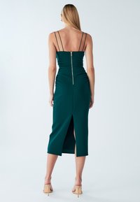 Šaty smaragdově zelené barvy s těsně přiléhajícím živůtkem, řasením po stranách a zipem na zádech. Mají tenká ramínka a rozparek sahající až ke stehnům.