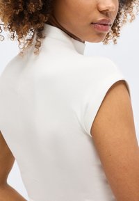 Κοντινό πλάνο γυναίκας με σγουρά μαλλιά που φοράει λευκό κοντομάνικο τοπ, δείχνοντας τον ώμο της και μερικό πρόσωπο, στραμμένο ελαφρώς προς τα έξω.