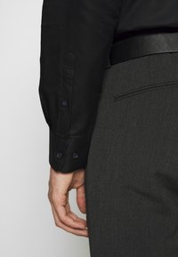 Camicia nera con bottoni e polsini, abbinata a pantaloni grigi scuri caratterizzati da una texture elegante e una cintura in pelle nera coordinata.
