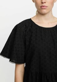 Sort øjeletkjole med korte, lagdelte ærmer med bølgede kanter, rund hals og et løst, samlet bodice-design med blomsterdekor.
