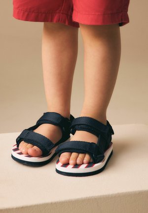 Jambes inférieures d'un enfant portant un short rouge et des sandales noires à velcro avec des semelles rayées, debout sur une surface claire.