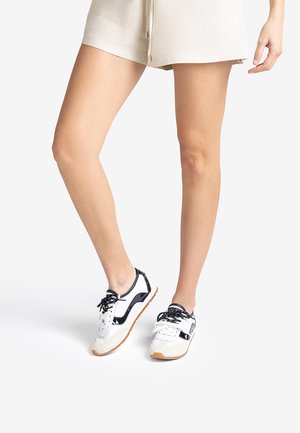 Woman wearing beige shorts and white sneakers with black details, standing against a plain white background.