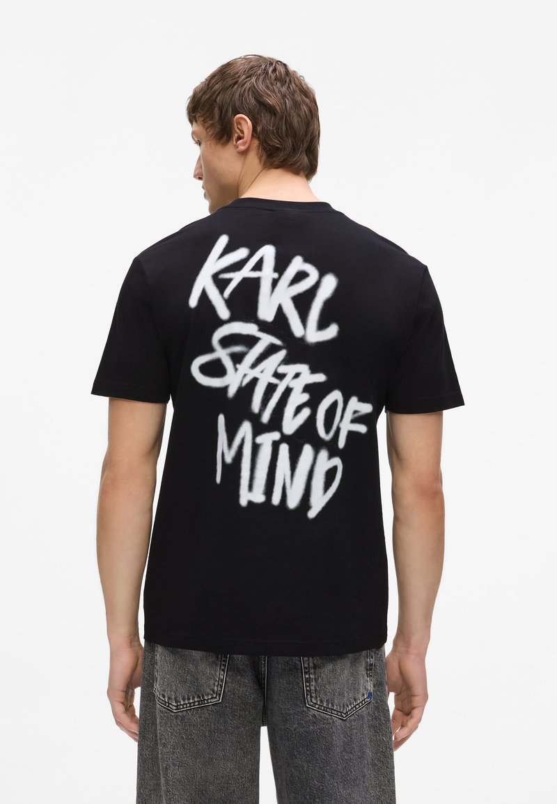 Άνδρας που φοράει μαύρο T-shirt με λευκό κείμενο σε στιλ γκράφιτι "KARL STATE OF MIND" στο πίσω μέρος, συνδυασμένο με γκρι σορτς από τζιν.
