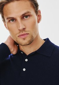 Granatowa dzianinowa koszulka polo z klasycznym kołnierzykiem, trzema białymi guzikami i strukturalną tkaniną. Model ma krótkie włosy i neutralny wyraz twarzy.