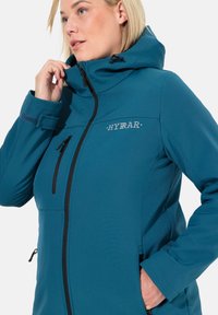 Giacca softshell turchese con design aderente, dotata di zip frontale, tasche laterali e cappuccio. Logo "HYRAR" sul petto.