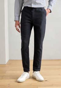 Dark navy pants made of a lightweight fabric feature an elastic waistband and side pockets, paired with white sneakers on a wooden floor.