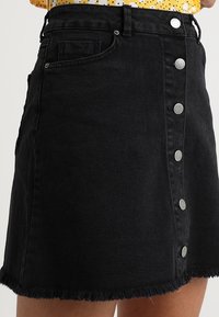 Schwarzer Denimrock mit ausgefranstem Saum, vorne mit fünf silbernen Knöpfen geschlossen und einer seitlichen Tasche.