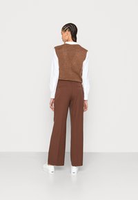 Colete castanho tricotado sobre uma camisa de manga longa branca, combinado com calças largas castanhas. Ténis brancos completam o visual.
