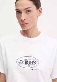 T-shirt bianco in cotone con una stampa grafica floreale che presenta la scritta "adidas" in nero e dettagli viola. Maniche corte e colletto a girocollo.