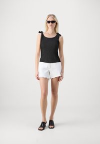 Haut sans manches noir avec des bretelles nouées, associé à un short blanc effiloché et des mules noires à bouts ouverts ; texture lisse et coupe moderne.