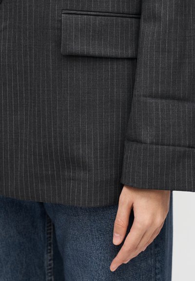 Blazer gris foncé à rayures fines avec un tissu texturé, doté d'une poche poitrine et d'une coupe décontractée. Porté sur un denim bleu.