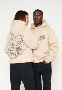 Sudaderas beige con impresiones de texto en la parte trasera. Una tiene "ESPÍRITU DE SOLIDARIDAD, COHESIÓN, UNIDAD", la otra presenta "MULTIPLY ORIGINALS EST. 2015."