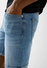 Shorts in denim azzurri con orlo arrotolato, cinque tasche e chiusura con bottone visibile. Texture leggermente sdrucita e dettagli di cucitura.