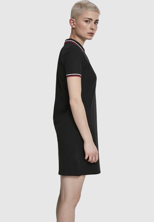 Personne aux cheveux blonds courts portant une robe noire à manches courtes avec des bordures rouges et blanches au col et aux poignets, vue de profil.