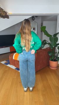 Άτομο με μακριά μαλλιά που φοράει πράσινο φούτερ και μπλε τζιν, στέκεται μέσα στο σπίτι μπροστά από έναν κόκκινο καναπέ και ένα μεγάλο γλαστρόφυτο.