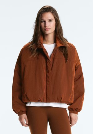 Žena s dlhými hnedými vlasmi, ktorá má na sebe púdrovú bundu tehlovej farby cez biele tričko a tehlové nohavice, otočená čelom.