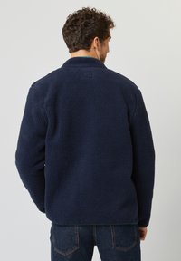 Navyblaue Fleecejacke mit einem strukturierten, plüschigen Äußeren, klassischem Kragen und einem quadratischen Logodetail auf dem oberen Rücken.