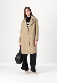 Beige, teksturert frakk med krage, knappelukking og sidelommer, brukt over en svart stripet genser og parret med løse svarte bukser.
