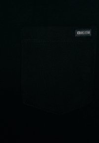 Juodo audinio artimas vaizdas, kuriame matyti siūtas kvadratinis kišenė su mažu „BIG STAR“ etikete viršutiniame dešiniajame kišenės kampe.