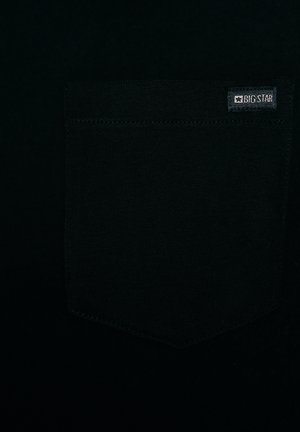 Detailní záběr černé látky se sešitou čtvercovou kapsou, na jejímž pravém horním rohu je malá etiketa s nápisem "BIG STAR".