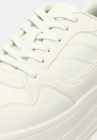 Baltas sportinis batelis, pagamintas iš lygaus sintetinės medžiagos, su plokščiomis raištelėmis, siuvinėtomis detalėmis ir storu, briaunuotu padu. Akcentai apima išlenktas šonines juosteles.