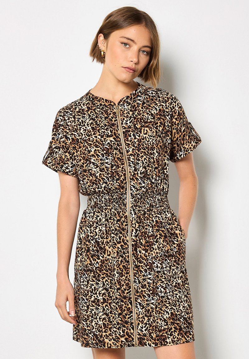 Femme aux cheveux courts portant une robe à manches courtes imprimé léopard avec une fermeture éclair à l'avant et une taille élastique, debout devant un fond uni.