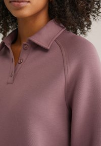 Polo majica u boji mauve s ovratnikom, s kopčanjem na gumbe i raglan rukavima, izrađena od glatkog tkanja sa blagim sjajem.